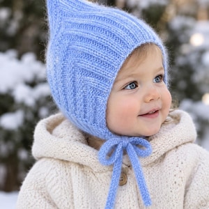 May include: A light blue knitted bonnet featuring a pointed top and a chevron-patterned texture. The bonnet is secured under the chin with two matching knitted ties that are tied into a bow. The knit fabric has a soft, fuzzy appearance.