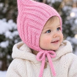 May include: A soft pink knitted bonnet featuring a pointed top and a chevron-style ribbed texture. The bonnet includes two knitted chin straps that tie into a bow under the chin, providing a secure fit with a fuzzy, textured yarn finish.