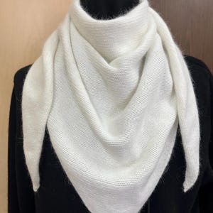 May include: A white, triangular knit scarf draped over a black garment. The scarf is made of a soft, textured material and is folded to create a layered effect. The scarf's ends point downward, creating a stylish and warm accessory.