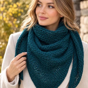 May include: A deep teal triangular shawl featuring a textured, open-knit stitch pattern. The shawl is draped around the neck and shoulders, showcasing a consistent, intricate weave throughout the fabric with a pointed bottom edge and tapered ends.