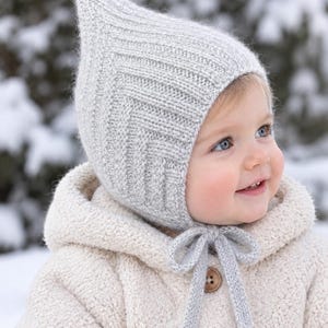 May include: A light gray knitted bonnet with a pointed top and a chevron-patterned texture. The bonnet features two matching knitted ties that are tied in a bow under the chin, providing a secure fit around the head and ears.