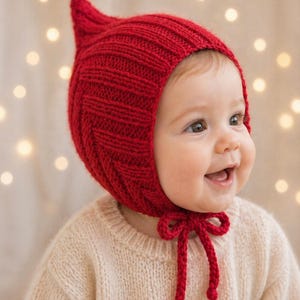 May include: A bright red knitted bonnet featuring a pointed top and a textured ribbed pattern. The bonnet includes two long knitted ties that fasten under the chin with a bow, providing a secure fit around the head with a distinct peak at the crown.