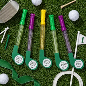 May include: A collection of golf-themed party favors, including bubble wands shaped like golf clubs in green, purple, and yellow. Each club has a green head and a label that reads "THANKS FORE SWINGING BY MY PAR-TEE NOAH". Golf balls and tees are also visible.