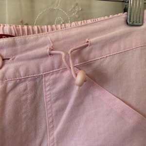 May include: Pink cotton pants with a drawstring waist and a button closure. The pants have a relaxed fit and a slightly cropped length.
