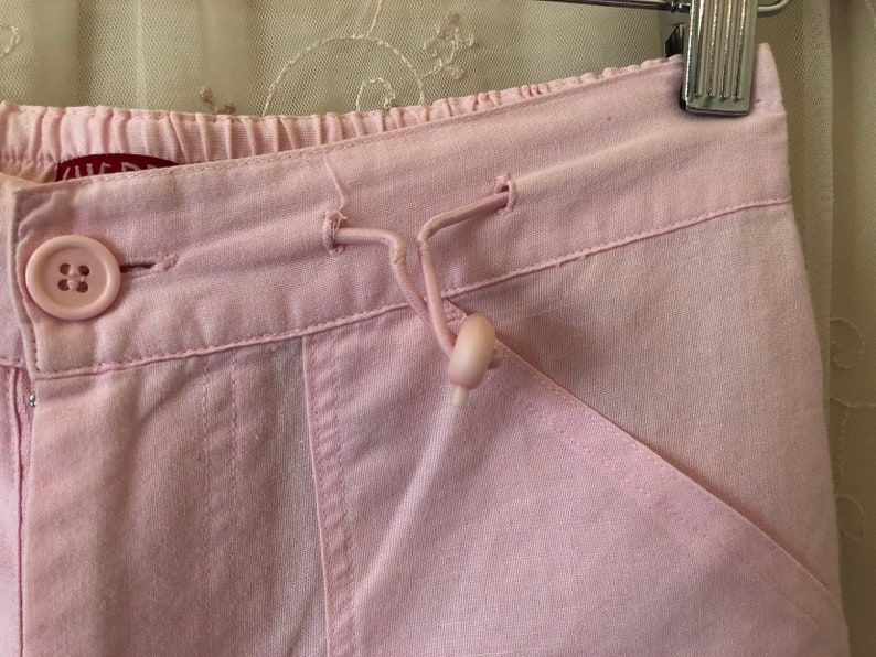 May include: Pink cotton pants with a drawstring waist and a button closure. The pants have a relaxed fit and a slightly cropped length.