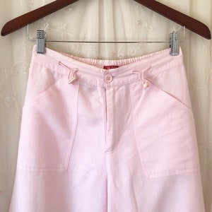 May include: Pink linen pants with a drawstring waist and two large pockets on the front. The pants have a button closure and a small red label that says "The Drop" on the front.