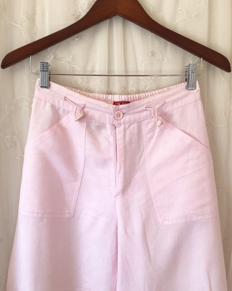 May include: Pink linen pants with a drawstring waist and two large pockets on the front. The pants have a button closure and a small red label that says "The Drop" on the front.