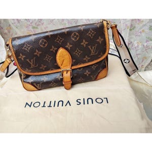 May include: Brown and tan shoulder bag with a buckle closure. The bag features a repeating pattern of brown logos and a tan leather trim. A white cloth with the words "LOUIS VUITTON" is beneath the bag.