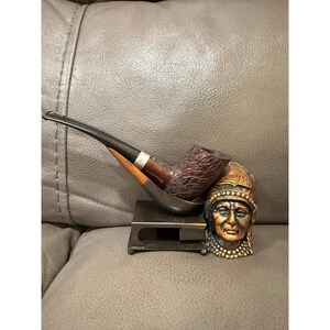 May include: A Peterson smoking pipe with a dark brown bowl and a black stem, resting on a metal stand. Beside it is a decorative Native American head figurine with a detailed headdress and facial features. The pipe and stand are on a grey surface.