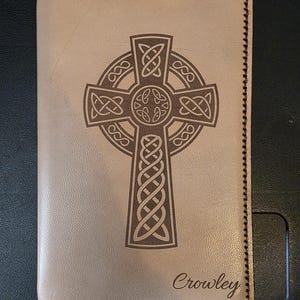 Könnte beinhalten: Ein hellbrauner Leder-Notizbuchumschlag mit einem keltischen Kreuzmuster in Braun. Das Wort "Crowley" ist in Schreibschrift unten geschrieben. Die Kanten sind mit dunkelbraunem Faden genäht.