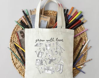 May include: Cream-colored tote bag with the phrase "grow with love" and a collection of gardening tools illustrated in black. The bag is surrounded by pencils and pens, and rests on a woven mat.