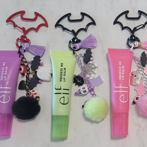 Op de afbeelding: Zeven e.l.f. Squeeze Me lippenbalsemtubes in verschillende kleuren, elk bevestigd aan een vleermuisvormige sleutelhanger. De sleutelhangers zijn voorzien van diverse bedels, waaronder kleine spookjes, schedels, vleermuizen en pompons.