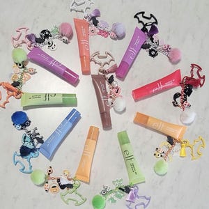 May include: A collection of ten e.l.f. Squeeze Me lip balm tubes in various colours, each attached to a decorative keychain featuring bat-shaped charms, colourful pom-poms, and small Halloween-themed trinkets like ghosts and pumpkins.