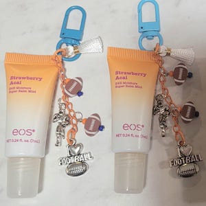 May include: Three lip balm tubes with football-themed keychains. The tubes are orange and white, with the text "Strawberry Acai" and "eos". Each keychain has a football charm, a football player charm, and a "FOOTBALL" charm.