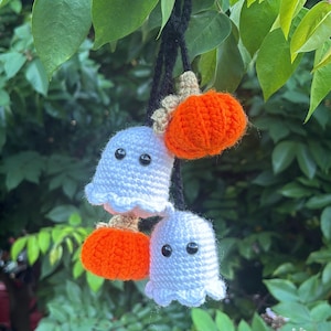 Puede incluir: Decoraciones de Halloween hechas a ganchillo. La imagen muestra dos fantasmas blancos con ojos de botón negros y dos calabazas naranjas, suspendidos de una cuerda negra. El fondo es follaje verde.