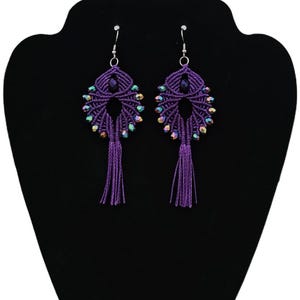 May include: A pair of handmade purple macrame earrings with iridescent beads and dangling tassels. The earrings are displayed on a black jewelry stand, showcasing their intricate woven design and vibrant color.