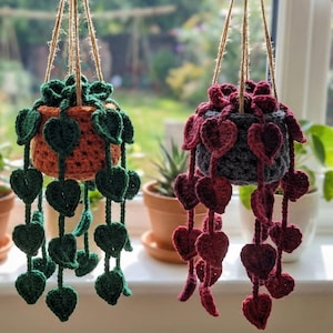 May include: Three hanging crochet plant holders in cream, orange, and burgundy, each with trailing leaf accents. The holders are suspended from a wooden branch by twine. The background features a window with greenery and potted plants.