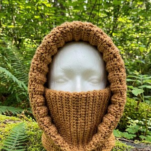 May include: A solid tan, crocheted balaclava featuring a thick, ribbed texture that covers the neck and frames the face. The headgear is constructed with a consistent yarn stitch pattern, creating a warm, unified appearance around the head.