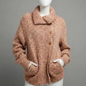 May include: A light brown, chunky knit sweater with a wide collar and two large pockets. The sweater has a button closure and is displayed on a mannequin. The knitwear has a speckled texture.