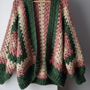 Peut inclure: Cardigan en crochet fait à la main, à devant ouvert, avec un motif de carrés granny. La palette de couleurs comprend des nuances de vert, de rose et de beige. Les manches sont longues et le cardigan est suspendu à un cintre en bois.