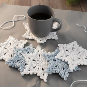 May include: A gray mug filled with dark liquid sits on a white snowflake-shaped coaster. Several crocheted snowflake coasters in white and light blue are arranged on a gray surface. White yarn is visible.