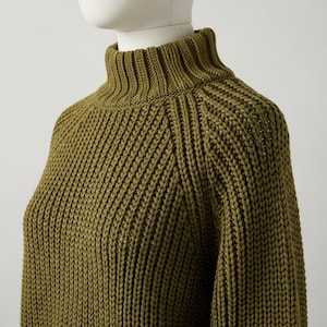 Puede incluir: Jersey de cuello alto verde oliva con diseño de punto acanalado. El jersey presenta un cuello alto y doblado y un patrón de punto diagonal texturizado en el cuerpo y las mangas. El primer plano resalta las costuras detalladas y el tono cálido y terroso del jersey.
