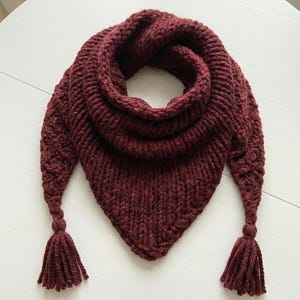 May include: A burgundy knitted scarf with a triangular shape and a cowl neck. The scarf has a thick, textured knit and features tassels at the ends. The scarf is displayed on a white surface.