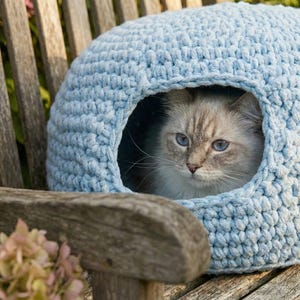 May include: A light blue, crocheted cat bed shaped like a dome, with a round opening. A fluffy, light-colored cat with blue eyes is inside the bed, resting on a wooden bench. The background includes a wooden bench and pink flowers.