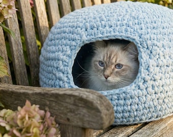 May include: A light blue, crocheted cat bed shaped like a dome, with a round opening. A fluffy, light-colored cat with blue eyes is inside the bed, resting on a wooden bench. The background includes a wooden bench and pink flowers.