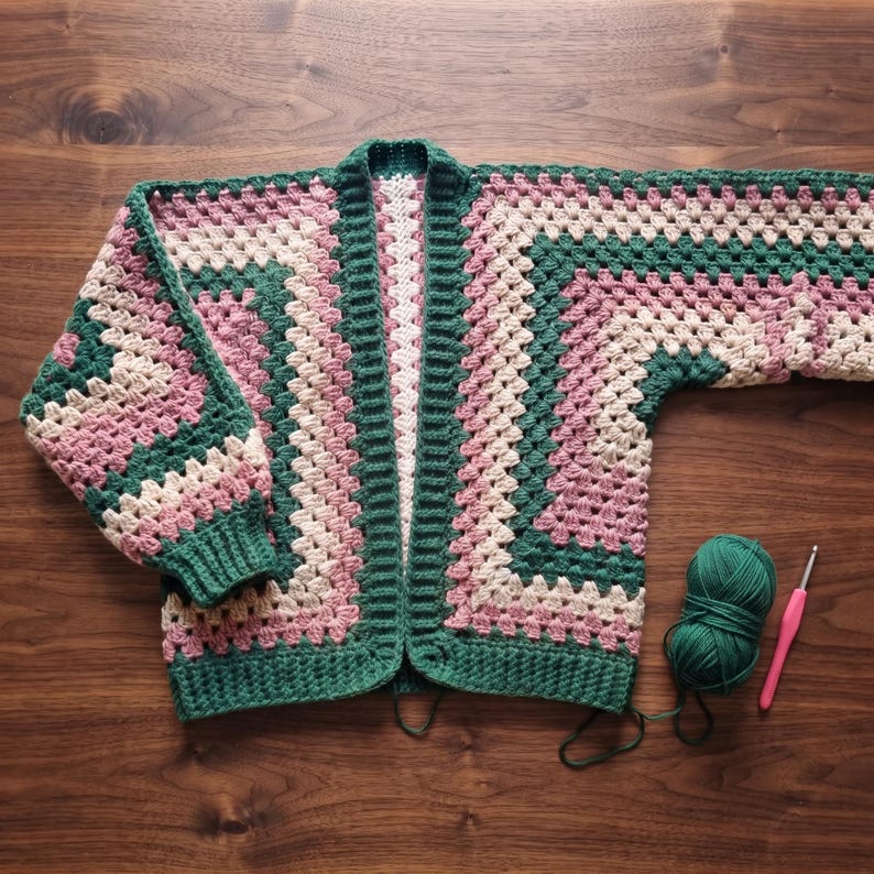 May include: A crochet cardigan featuring a granny square pattern in alternating bands of dark green, dusty pink, and cream yarn. The garment has long sleeves with ribbed cuffs, a ribbed front opening, and a matching ball of green yarn with a crochet hook.