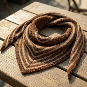 May include: A brown and beige striped knitted shawl, folded on a wooden surface. The shawl is triangular, with alternating stripes of light beige and dark brown. The knitted texture is visible, and the shawl appears soft and warm.