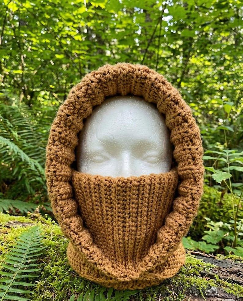 May include: A solid tan, crocheted balaclava featuring a thick, ribbed texture that covers the neck and frames the face. The headgear is constructed with a consistent yarn stitch pattern, creating a warm, unified appearance around the head.