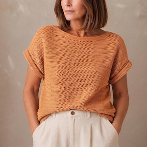 May include: A short-sleeved, burnt orange crochet top featuring a wide boat neckline and cuffed sleeves, paired with off-white trousers that have a visible button closure at the waist. The top has a consistent, textured knit pattern throughout.