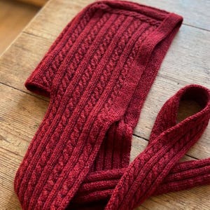 May include: A deep red knitted hood with an attached scarf. The hood and scarf are patterned with a cable knit design. The item is made from a soft, warm material, ideal for colder weather. The hood is on a wooden surface.
