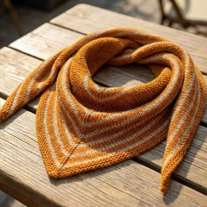 Puede incluir: Un chal triangular tejido a mano en tonos naranja y crema. El chal presenta un patrón de rayas y está drapeado sobre una superficie de madera. La cálida paleta de colores y el tejido texturizado sugieren un accesorio acogedor y hecho a mano.