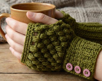 May include: Olive green crochet fingerless gloves with a textured pattern and three pink buttons. The gloves are worn while holding a brown mug. The background includes a knitted blanket and a wooden surface.