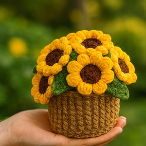 May include: A handcrafted crochet sunflower arrangement in a brown basket. The sunflowers have yellow petals and brown centers, with green leaves. The basket is a light brown color. The arrangement is held in a hand.