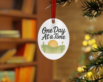 May include: White oval ornament with the text "One Day At a Time" in black script. The ornament features a watercolor design of a yellow sun setting over green hills with small yellow flowers. A red ribbon is attached for hanging.