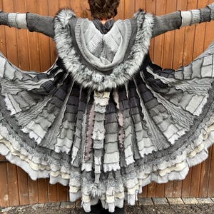 May include: A long, flared coat in shades of gray, charcoal, and white, featuring a hood trimmed with faux fur. The garment is constructed from multiple layered fabric panels with varied textures, including ribbed knit and frayed edges, and includes a long, dangling fabric tail.