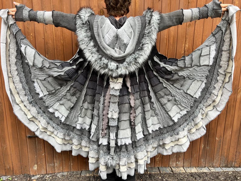 Peut inclure: Un long manteau &eacute;vas&eacute; dans des tons de gris, anthracite et blanc, dot&eacute; d'une capuche bord&eacute;e de fausse fourrure. Le v&ecirc;tement est compos&eacute; de multiples panneaux de tissu superpos&eacute;s aux textures vari&eacute;es, incluant du tricot c&ocirc;tel&eacute; et des bords effiloch&eacute;s, avec une longue queue en tissu.