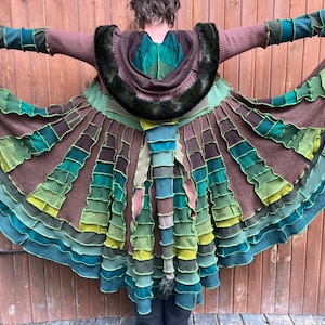 May include: A long, flared coat featuring a patchwork design of brown, teal, and lime green fabric panels. The garment includes a hood trimmed with dark faux fur, long sleeves with ribbed cuffs, and a long, segmented tail-like attachment at the back.