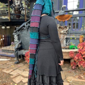 May include: A colorful, handmade knit hood with a long, layered tail. The hood features shades of teal, purple, and black. The person is wearing a black long-sleeved top and a tiered black skirt. The outfit is perfect for a costume or unique fashion statement.