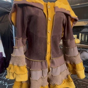 May include: A hooded jacket featuring a mustard yellow and dark brown color palette. The garment has a button-down front, long sleeves with tiered, ruffled cuffs, and a mix of textured fabrics including knit and suede-like panels throughout.