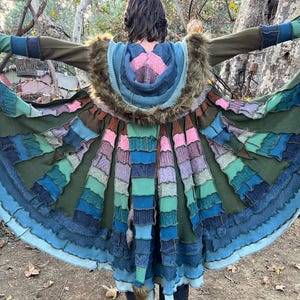 May include: A long, flared coat featuring a patchwork design of various knit and fabric textures in shades of olive green, blue, teal, and pink. The garment includes a hood trimmed with brown faux fur, long sleeves with fingerless glove cuffs, and a wide, circular hemline constructed from multiple layered, ruffled fabric panels.