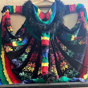 May include: A sleeveless garment constructed from black fabric panels interspersed with vibrant, multi-colored knitted sections featuring star patterns in red, yellow, green, and blue. The neckline and hem are trimmed with black faux fur fringe.