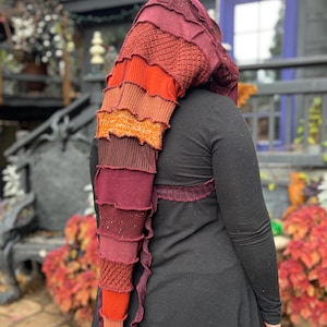 May include: A multi-toned, layered knit hood in shades of burgundy, orange, and gold. The hood is worn over a black long-sleeved top. The design features a cascading, textured pattern, creating a unique and artistic accessory.