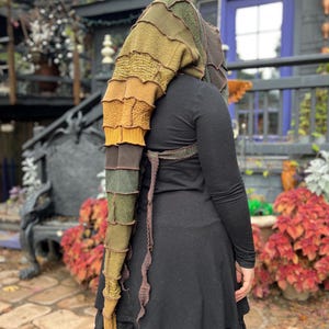 May include: A multi-toned, knitted hood in shades of green, brown, and gold. The hood is long and pointed, with a textured design. The person is wearing a black dress.