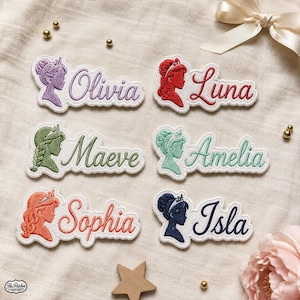 Op de afbeelding: Zes geborduurde naamemblemen met silhouetten van figuren met kronen. De namen Olivia, Luna, Maeve, Amelia, Sophia en Isla zijn geborduurd in bijpassende draadkleuren, waaronder paars, rood, groen, taling, oranje en donkerblauw.