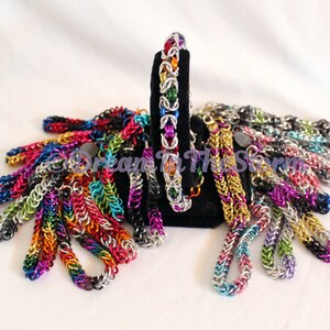 Puede incluir: Una colección de pulseras de malla de cadena de colores en varios colores, incluyendo plata, oro, negro, rojo, verde, azul y morado. Las pulseras se muestran en un soporte de terciopelo negro.