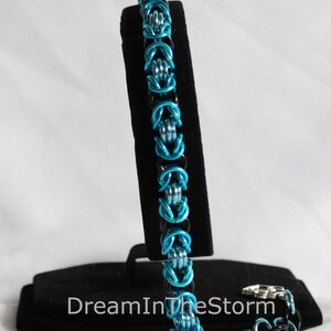 Puede incluir: Un brazalete de malla de cadena azul y negro con acabado plateado. El brazalete se muestra en un soporte negro con el texto "DreamInTheStorm".
